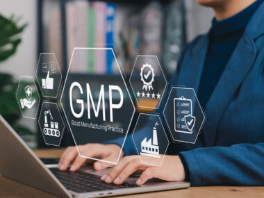 Icons for quality control and good manufacturing practices are seen above the hands of a person typing on a laptop keyboard.