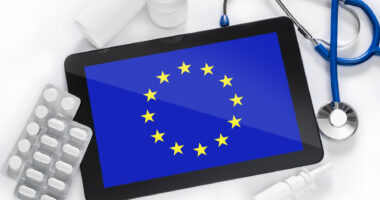 A tablet with an image of the European Union flag is surrounded by prescription medications and a stethoscope.