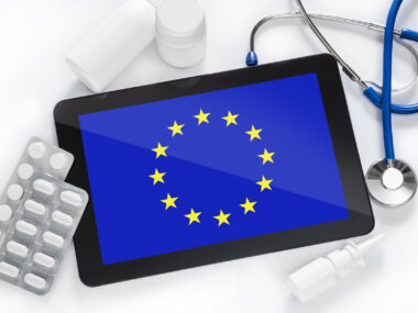 A tablet with an image of the European Union flag is surrounded by prescription medications and a stethoscope.