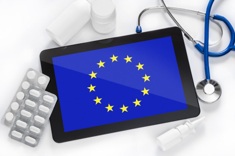 A tablet with an image of the European Union flag is surrounded by prescription medications and a stethoscope.