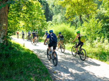 A group of cyclists rides along a trail surrounded by trees.