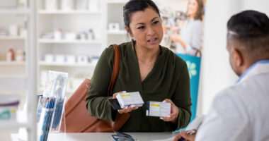 A drugstore customer, seen holding two medication boxes, consults with a pharmacist.