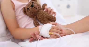 An adult holds the bandaged hand of a child in a hospital bed receiving an infusion, while the youngster clutches a teddy bear with the other hand.