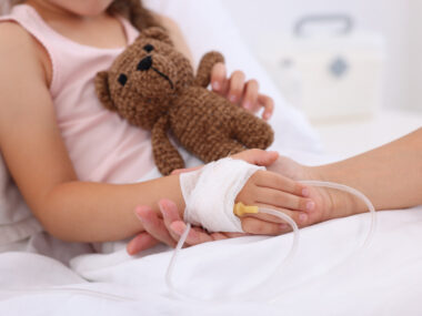 An adult holds the bandaged hand of a child in a hospital bed receiving an infusion, while the youngster clutches a teddy bear with the other hand.