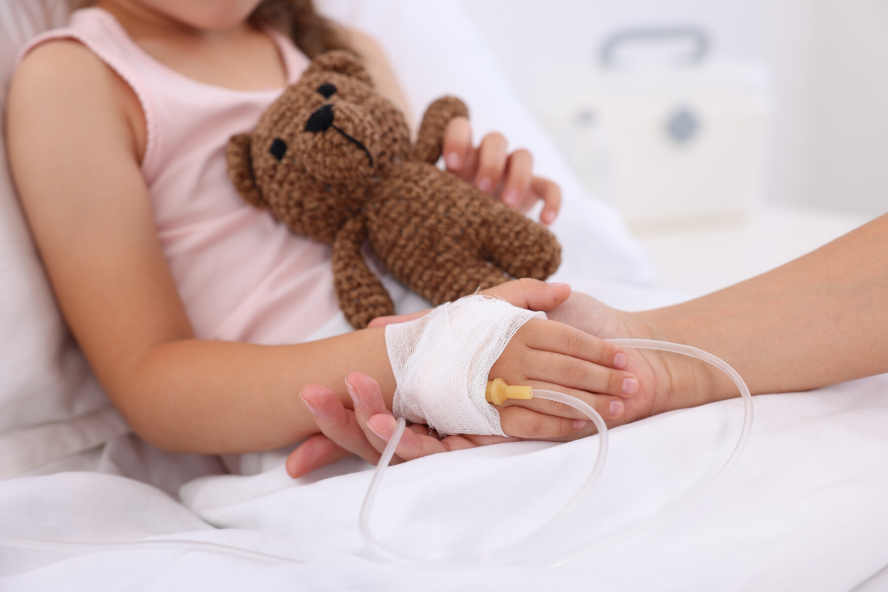 An adult holds the bandaged hand of a child in a hospital bed receiving an infusion, while the youngster clutches a teddy bear with the other hand.