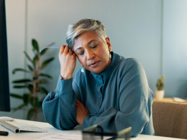 A tired-looking woman is shown seated at a table with her hand on the side of her head.