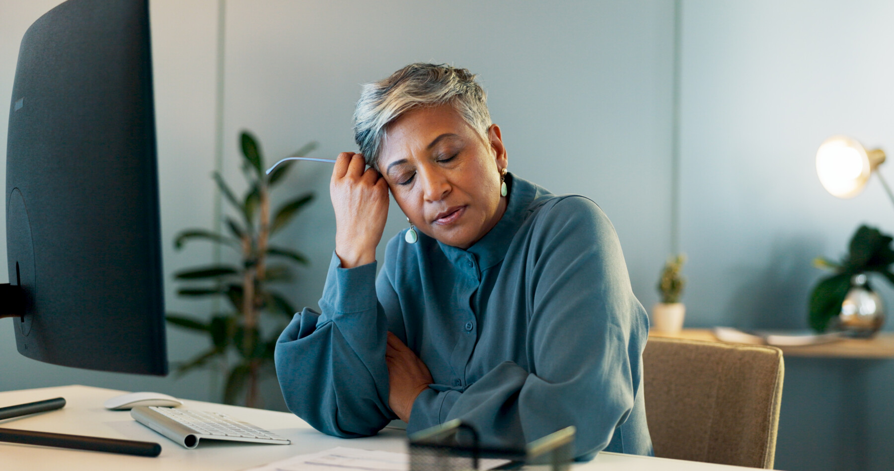 A tired-looking woman is shown seated at a table with her hand on the side of her head.