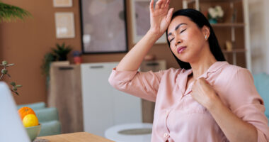 A woman experiencing a hot flash and headache during menopause holds her hand to her forehead while working on her laptop.