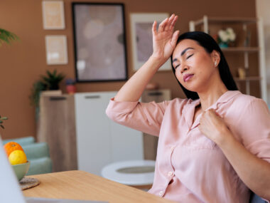 A woman experiencing a hot flash and headache during menopause holds her hand to her forehead while working on her laptop.