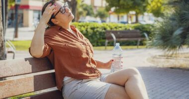 A woman sits on a park bench with one hand to her head and another holding a water bottle.