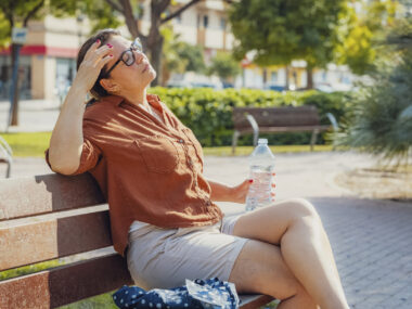 A woman sits on a park bench with one hand to her head and another holding a water bottle.