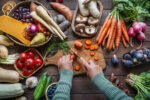 Overhead view of a person slicing carrots on a wooden cutting board surrounded by fresh vegetables and fruit.