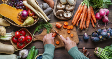 Overhead view of a person slicing carrots on a wooden cutting board surrounded by fresh vegetables and fruit.