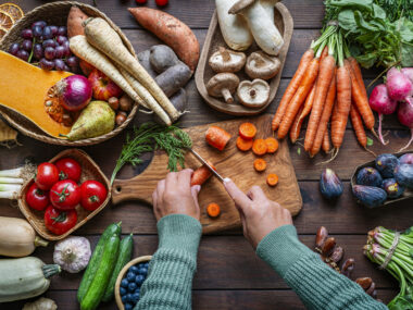 Overhead view of a person slicing carrots on a wooden cutting board surrounded by fresh vegetables and fruit.