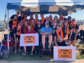 A large group of people huddle under a canopy for a photo on a sunny day. The picture is blurry, but it seems they're all wearing orange leis. Some of the people in the front of the photo are in wheelchairs. 