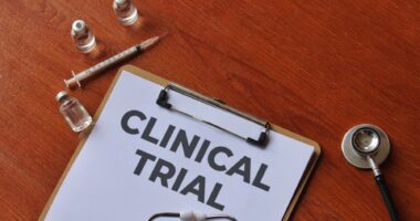 A clipboard with the words CLINICAL TRIAL on a wooden table along with a syringe, vials, and a stethoscope.