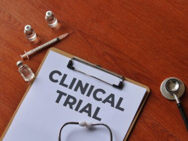 A clipboard with the words CLINICAL TRIAL on a wooden table along with a syringe, vials, and a stethoscope.