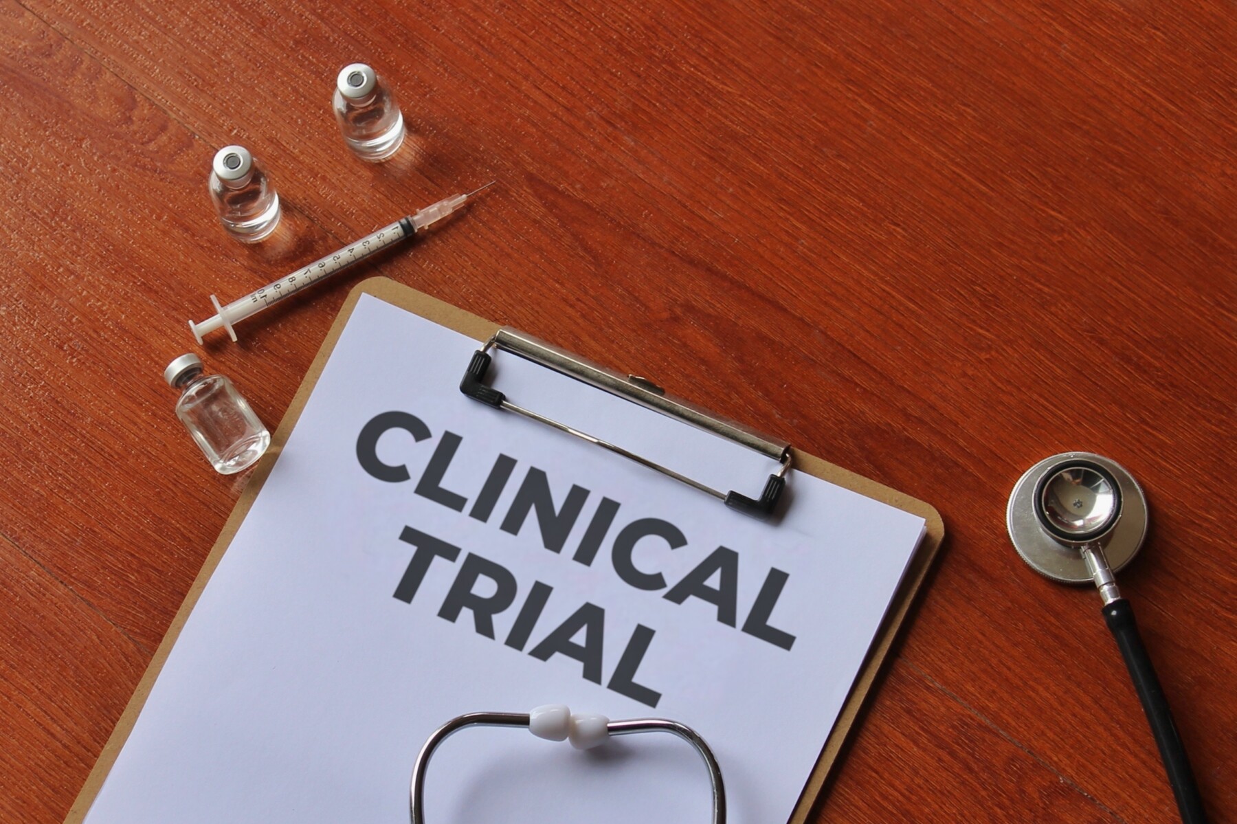 A clipboard with the words CLINICAL TRIAL on a wooden table along with a syringe, vials, and a stethoscope.