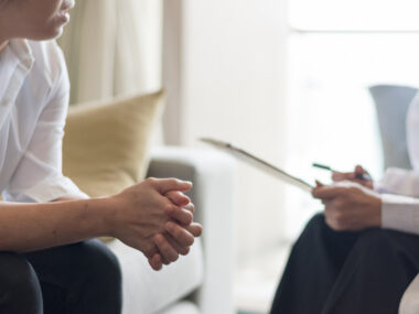 A physician holds a tablet while conferring with a patient, who sits with their hands clasped together.