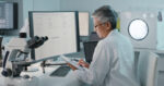 A researcher seated at a desk with a computer and microscope reads data on a tablet.