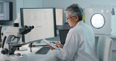 A researcher seated at a desk with a computer and microscope reads data on a tablet.