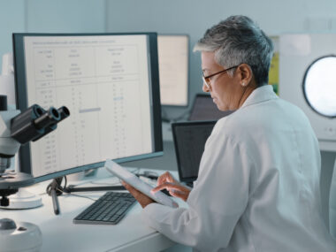 A researcher seated at a desk with a computer and microscope reads data on a tablet.