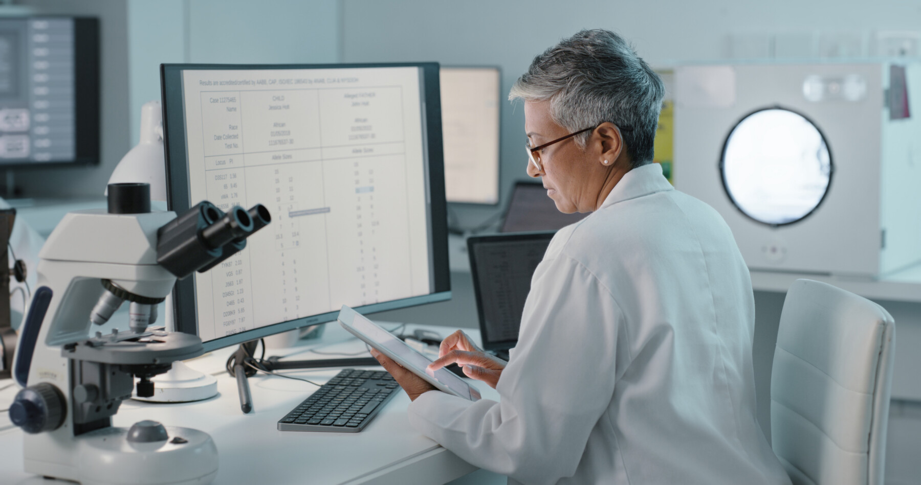 A researcher seated at a desk with a computer and microscope reads data on a tablet.
