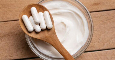 A wooden spoon with white capsules rests over a bowl of yogurt.