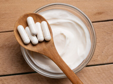 A wooden spoon with white capsules rests over a bowl of yogurt.