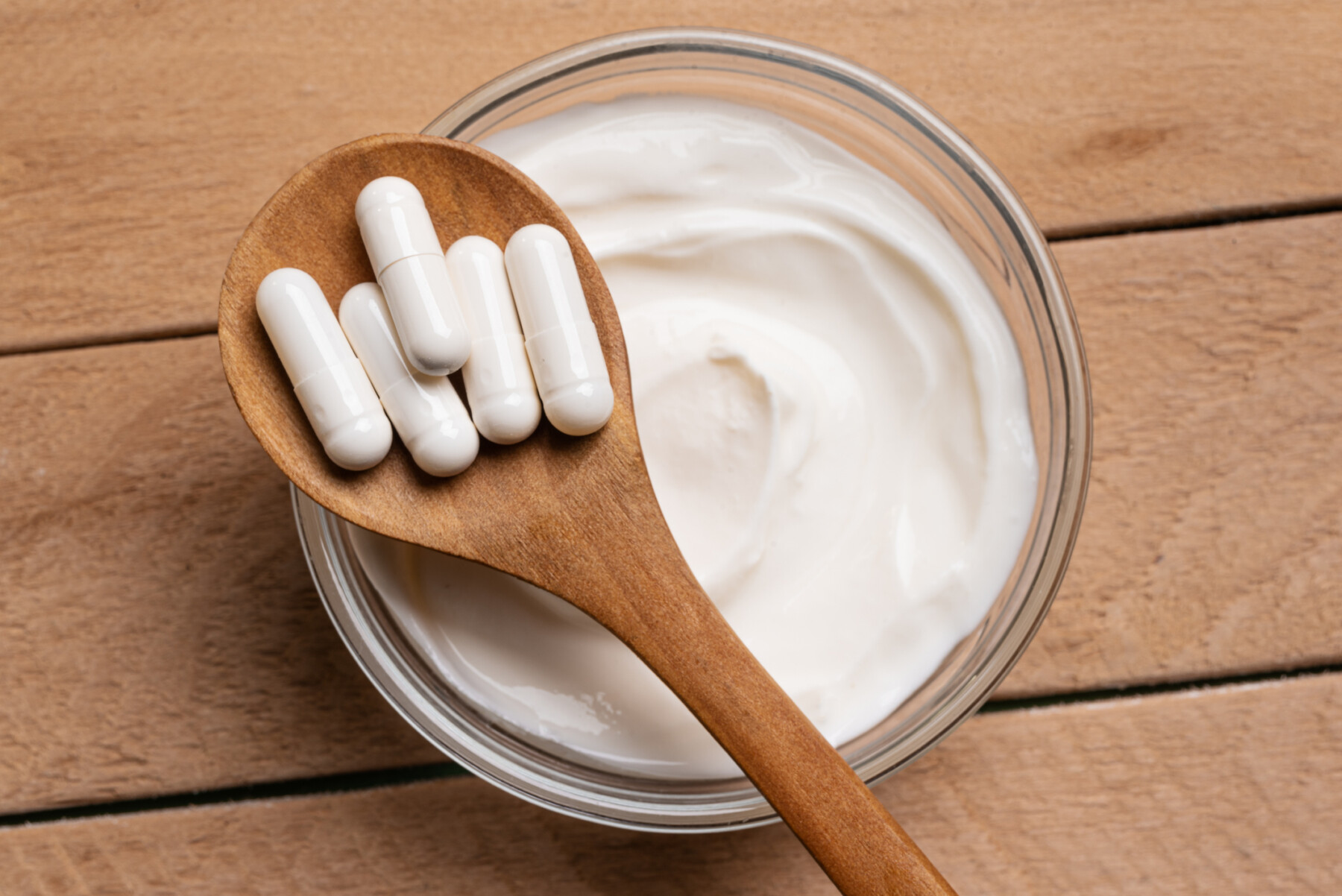 A wooden spoon with white capsules rests over a bowl of yogurt.