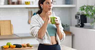 A woman stands in a kitchen drinking a green smoothie.