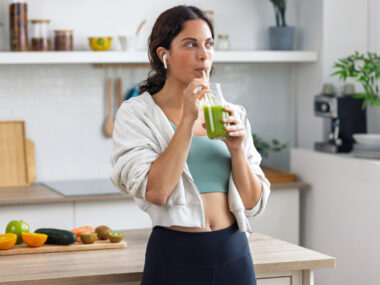 A woman stands in a kitchen drinking a green smoothie.