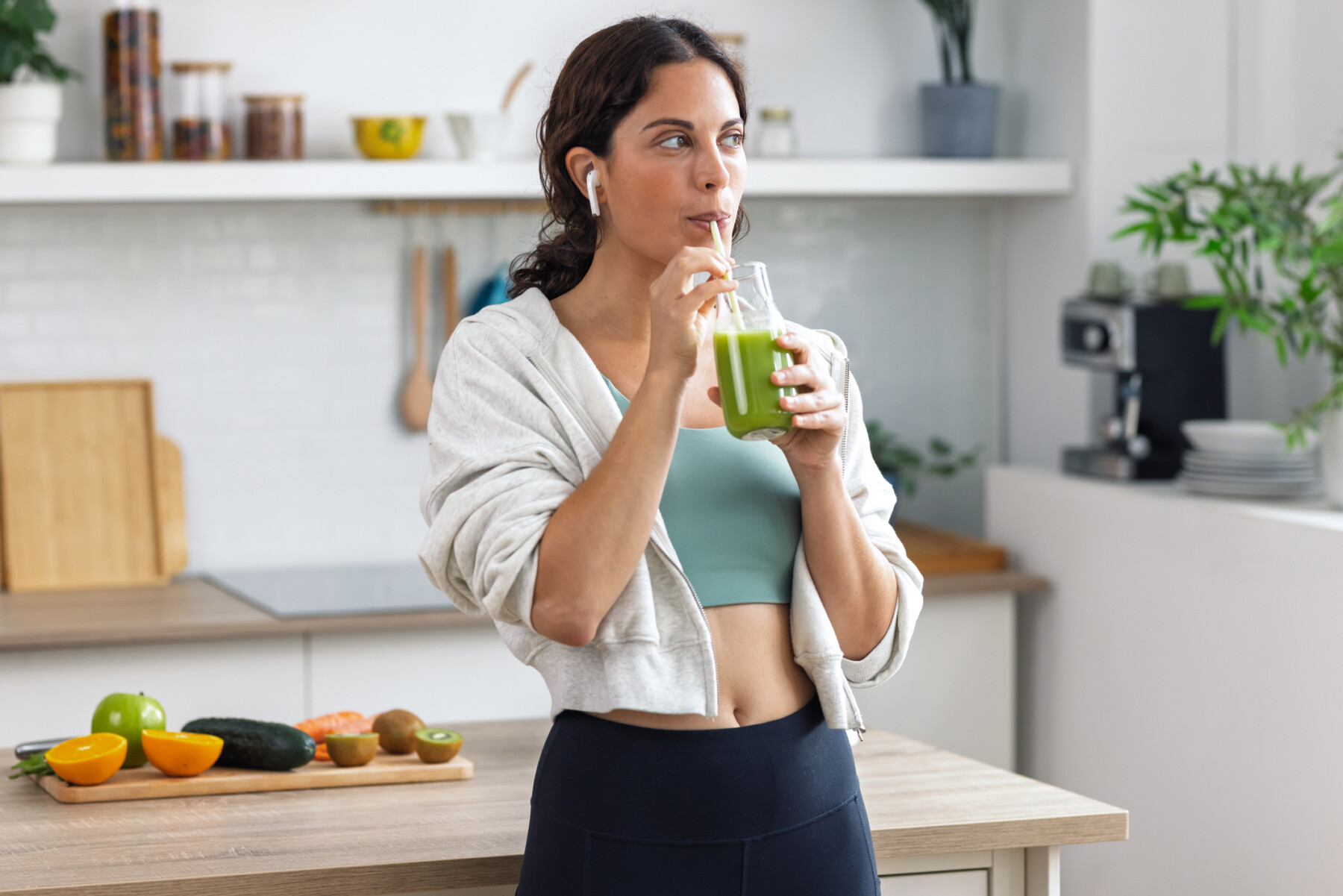 A woman stands in a kitchen drinking a green smoothie.