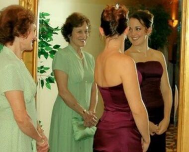 Two women stand before a mirror, through which they glance at each other.