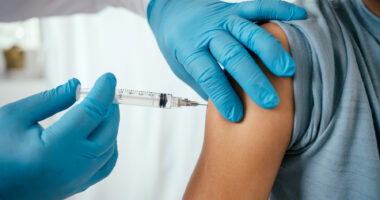 A close-up image shows gloved hands injecting a vaccination needle into a person's upper arm.