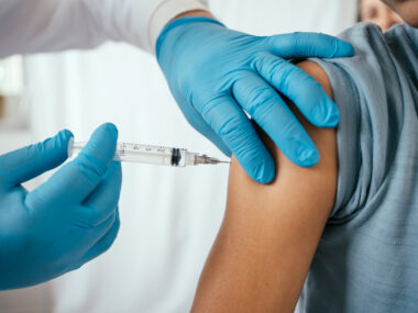 A close-up image shows gloved hands injecting a vaccination needle into a person's upper arm.