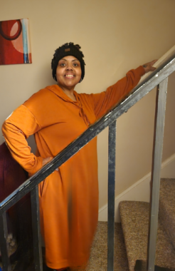 A woman wearing an orange dress stands on a carpeted staircase, with her left hand on a railing and her right hand on her hip. She's angled toward the camera to her right and is smiling proudly.