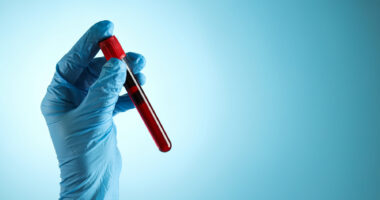 A blue-gloved hand holds a test tube filled with blood.
