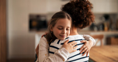 A young girl hugs a parent in a home kitchen.