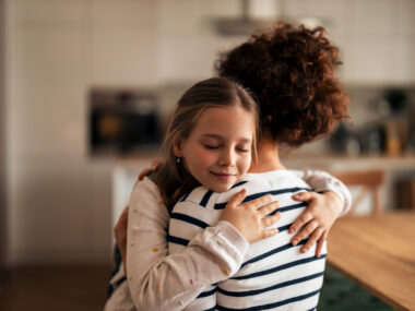 A young girl hugs a parent in a home kitchen.