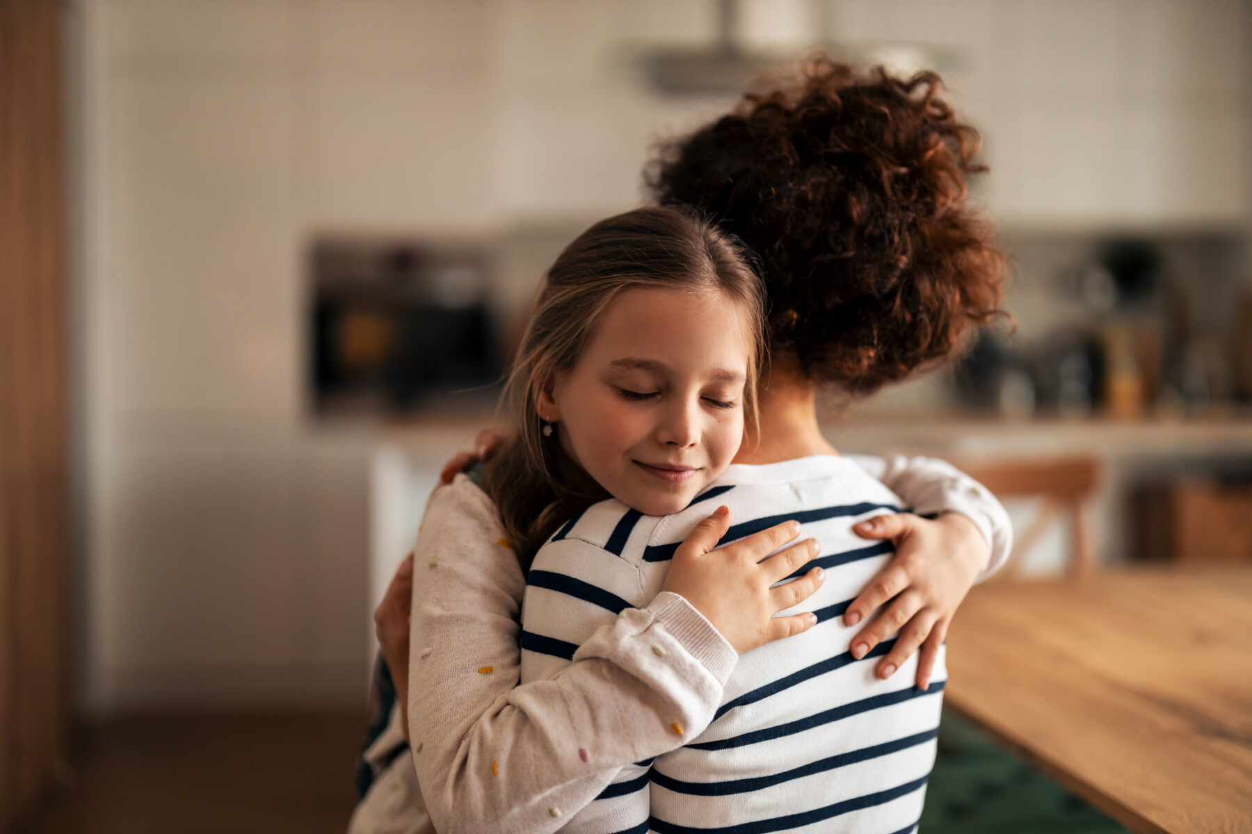 A young girl hugs a parent in a home kitchen.