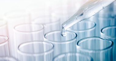 Close-up of a pipette dropping clear liquid into glass test tubes.