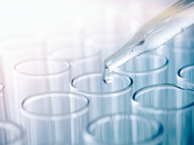 Close-up of a pipette dropping clear liquid into glass test tubes.