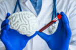 A person in a white coat with blue gloves holds a model of a brain in one hand and a blood-filled test tube in the other.