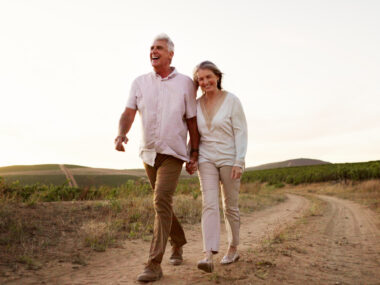 A middle-aged man and woman smile and hold hands as they walk along a dirt road in the countryside.