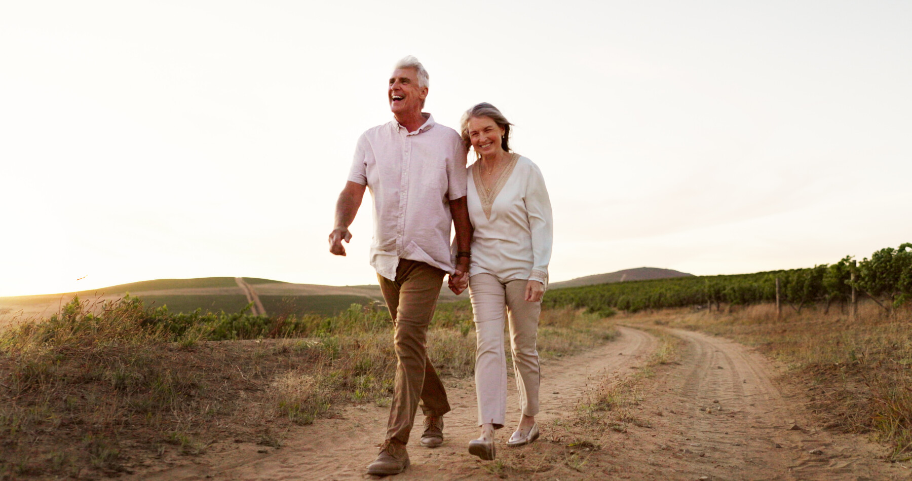 A middle-aged man and woman smile and hold hands as they walk along a dirt road in the countryside.