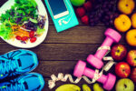 Healthy food, including a salad and fresh fruits, and exercise equipment including running shoes, a smartphone, and dumbbells, are arranged on wood background.