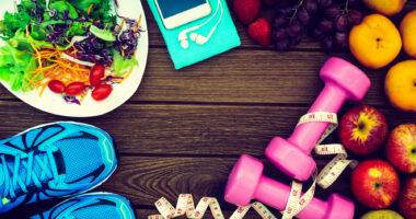 Healthy food, including a salad and fresh fruits, and exercise equipment including running shoes, a smartphone, and dumbbells, are arranged on wood background.