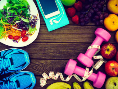 Healthy food, including a salad and fresh fruits, and exercise equipment including running shoes, a smartphone, and dumbbells, are arranged on wood background.