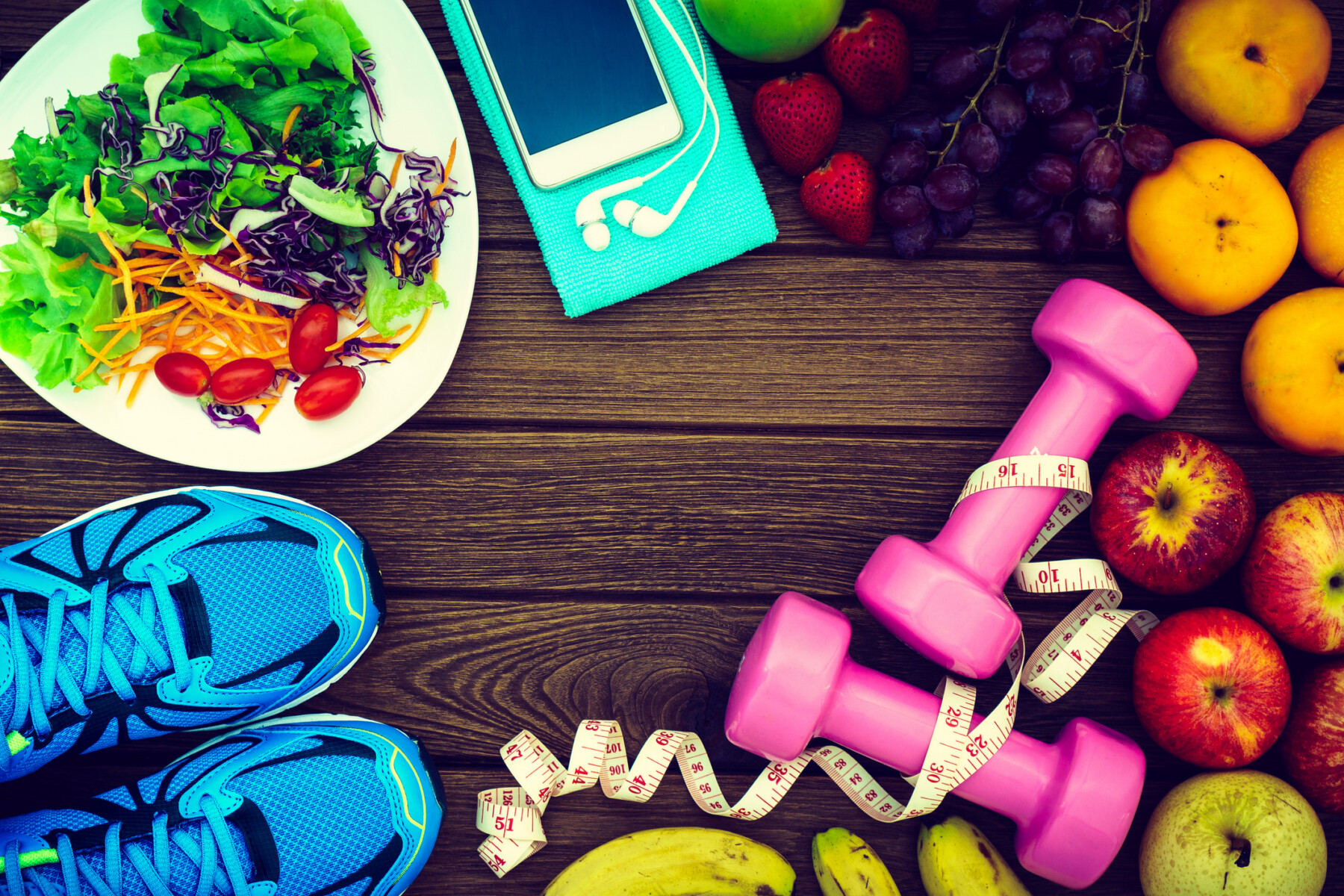 Healthy food, including a salad and fresh fruits, and exercise equipment including running shoes, a smartphone, and dumbbells, are arranged on wood background.